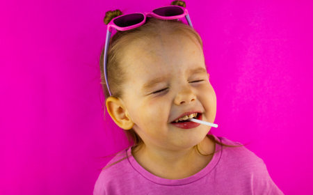 Beautiful cute baby girl screwed up her eyes with a lollipop in her mouth on a pink background. The child smiles happily in a candy. The concept of a cute baby screwed up his eyes with a lollipop.の写真素材