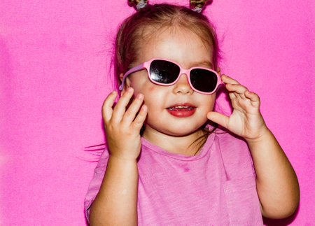 Humor. On a pink background, a little girl child funny adjusts her sunglasses that have fallen off. The child is funny trying to return the sunglasses that have fallen into place.の写真素材