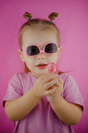 A cute child in sunglasses holds a lollipop in his hands on a pink background. Little girl child with a lollipop posing stylishly on a pink background. Concept of cute baby with lollipop.の写真素材
