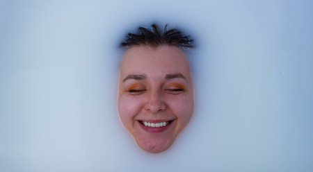 A womans face immersed in anu with milk with open eyes and smiling. A woman smiles sweetly while lying in a bath filled to the brim with milk. Body care concept.の写真素材