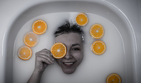 A beautiful woman lies in a bath with milk and bright juicy oranges and smiles broadly while looking at the camera A woman is holding a circle of orange while lying in a bath of milk. View from above.の写真素材
