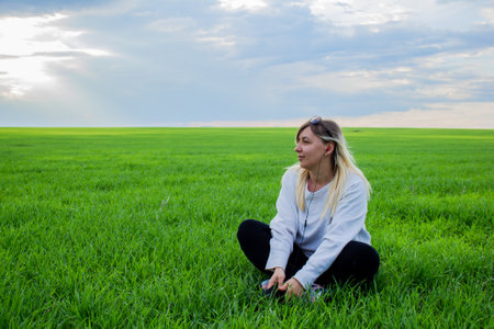 A beautiful woman listens to music in headphones while sitting on a green field against a cloudy sky. Pretty woman looks away thoughtfully and listens to music on headphones.の写真素材
