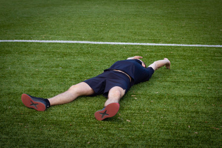 A man lies resting on a sports football field in a stadium. A man is lying on the green lawn of a sports field for playing football. A man is resting after an active football match on the field.の写真素材