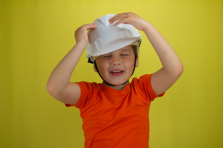 Child boy frustratedly grabbed the construction helmet on his head on a yellow background. Isolated....