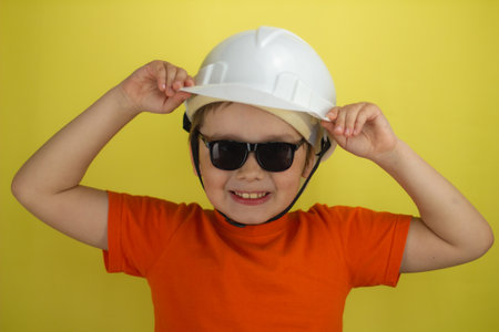 In sunglasses, a boy adjusts a construction helmet on a yellow background and smiles. Child boy engineer millo smiling in stylish sunglasses and construction helmet. Child engineer.の写真素材