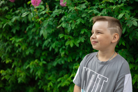 In summer, a child in a t-shirt boy stands against the background of green bushes. Copy space. Portrait of a child looking away with a wistful smile. Concept boy portrait on greenery background.の写真素材