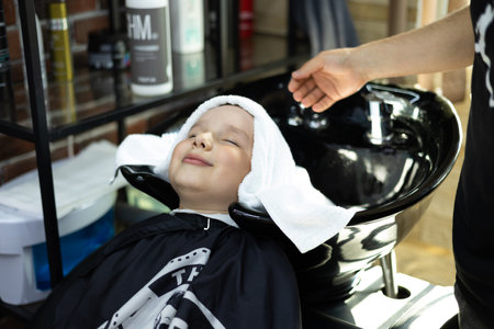 The boy closed his eyes and enjoys while the hairdresser gently wipes his head with a towel in a beauty salon. After washing the hair in the barbershop, the child wipes his head with a fluffy towel.の写真素材