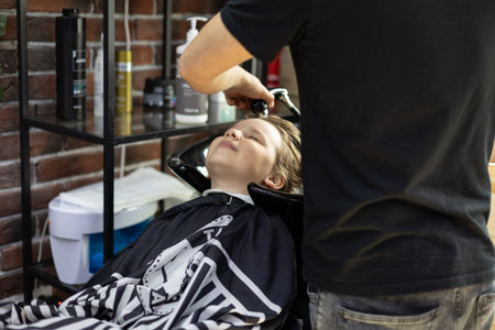To a child boy, a hairdresser washes his head in the sink before a haircut. The boy came to the barbershop for a haircut. The hairdressers hands gently wash the childs head in the sink.の写真素材