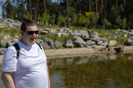 On a sunny summer day, a man with a backpack and sunglasses is resting on the shore of a beautiful lake in the mountains. A traveler in a white T-shirt stands near a mountain lake in summer.の写真素材