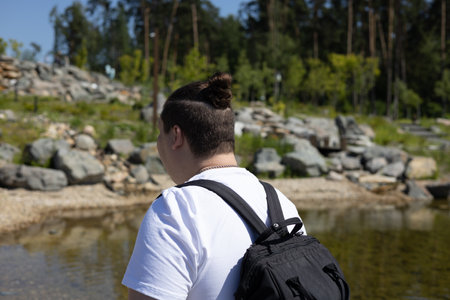 A male traveler with a backpack stands with his back to the camera against the backdrop of a mountain pond strewn with stones. In summer, a man travels through a mountain park.の写真素材