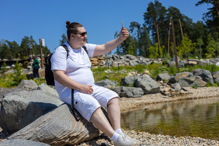 On a summer sunny day, a man sits on a stone by the lake and takes a selfie on the phone. A man is photographed for memory on his phone. Summer selfie travel concept.の写真素材