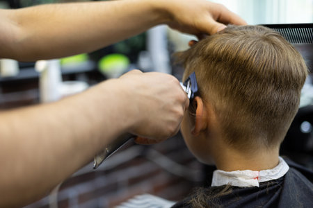 View from the back. The hairdresser cuts the hair on the boys head with a machine in a beauty salon. The child calmly gives the hairdresser a haircut.の写真素材