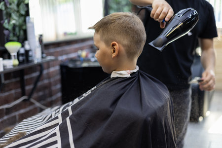 With the help of a hair dryer, the parkmacher blows the cut hair from the neck and shoulders of the boys child. The hairdresser makes a fashionable haircut for the child for the summer.の写真素材