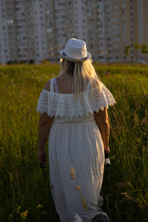 View from the back. A woman in a white sarafen walks home on a green meadow at sunset. The suns rays beautifully illuminate the silhouette of a woman with a golden light.の写真素材