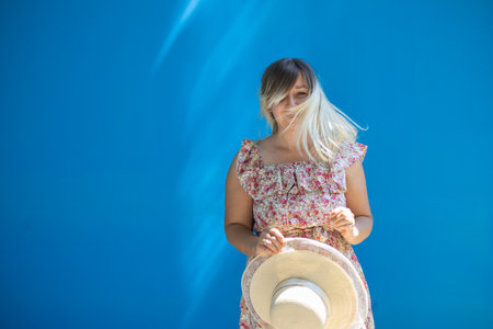 Copy space. Cute woman in summer on a blue background with flying hair and sun rays on her face, smiling at the camera. A beautiful woman in a coat with flowers smiles sweetly.の写真素材