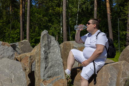 After a long journey, a man drinks water from a bottle, sitting on a large stone at the edge of the forest. A handsome traveler rests while sitting on a boulder with a backpack on his shoulders.の写真素材