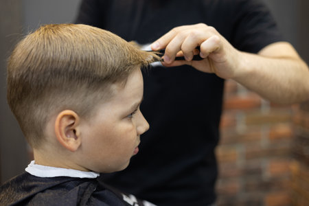 A boys child is being cut by a professional hairdresser. The boy sits in a chair with a cape on his shoulders and watches the work of a hairdresser. The concept of a child in a beauty salon.の写真素材