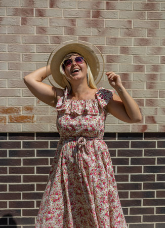 vertically. Woman on the background of a brick city wall in sunny weather. A beautiful woman in a srafan and a hat laughs against the backdrop of a brick wall. Conceptual summer portrait in the city.の写真素材