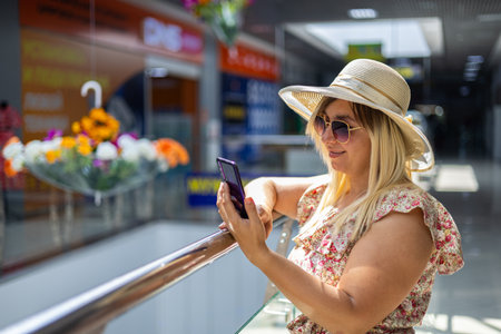 Portrait of a beautiful woman in a shopping mall with a mobile phone. A woman stands near the railing in a shopping center in a summer sundress and writes a message on her phone.の写真素材
