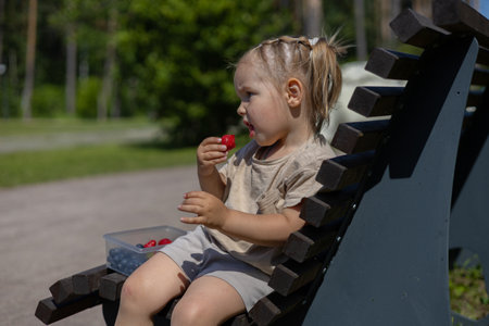 On a wooden bench in the park, a child eats strawberries in the summer. A girl in the summer heat enjoying ripe fragrant strawberries. The concept of a healthy summer snack with berries.の写真素材