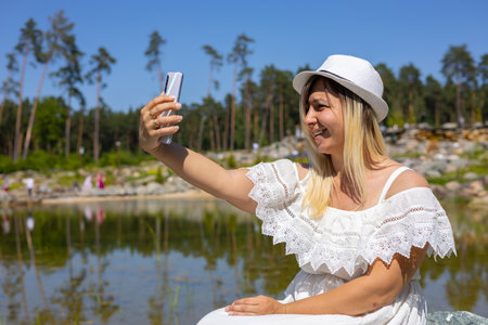 In summer, a woman makes a sefie on the phone on the shore of a beautiful lake in the park. The woman sat down to rest on a stone by the lake and decided to take a selfie as a keepsake.の写真素材