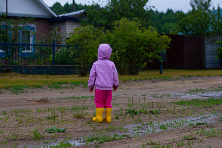 A child walks along a village road after the rain. A little girl in a pink jacket and yellow rubber boots walks along the ridge after joj. The child enjoys a happy childhood.の写真素材