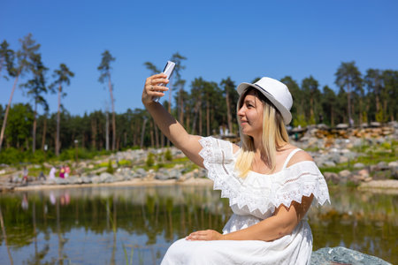 A cute woman takes a selfie on her phone while sitting on the shore of a beautiful lake. On a summer day, a woman in a dress is photographed on the phone against the backdrop of the lake.の写真素材
