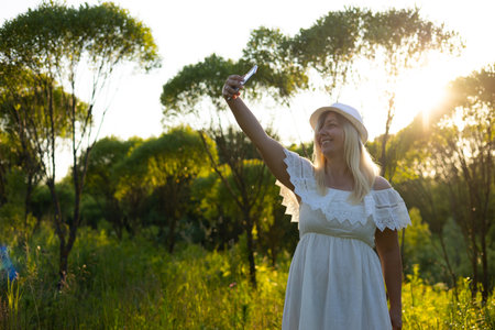 In the summer, in the rays of the setting sun, a cute woman in a white sundress takes a selfie on her phone. A woman takes a selfie against the background of beautiful lush greenery in the park.の写真素材