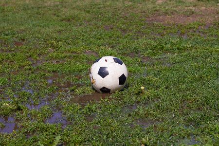 A soccer ball lies on the grass among puddles and ridges in summer. There is a ball in a puddle on the football field. In the summer on the lawn licks a soccer ball without people.の写真素材