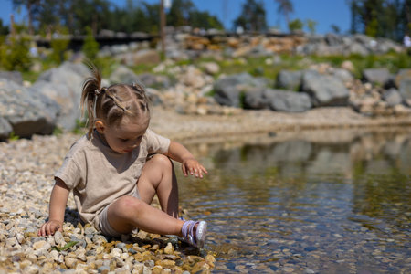 A child splashes in the water sitting on the rocky shore of the lake in summer. A beautiful little girl enjoys summer days on the shore of a picturesque lake. The concept of vacation near the water.の写真素材