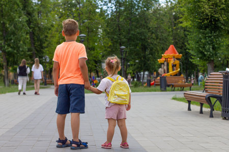 View from the back. Two children walk in the park holding hands in the summer. In shorts and T-shirts, children go to the childrens playground through the green park. Summer vacation concept.の写真素材