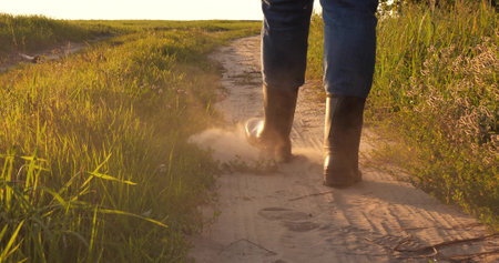 A farmer in rubber boots walks along a dusty road along a field. Mens feet tread on a harvested field at sunset. Raising the dust, the peasant walks along the path at sunset.の写真素材