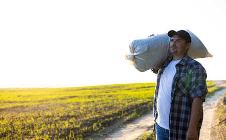 Wiping the sweat from his forehead with his hand, the farmer carries the harvested crop in a bag on his shoulder. Against the backdrop of a blue sky and a farmer man carries the harvest.の写真素材