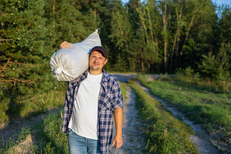 The farmer is holding a bag of hay on his shoulder and looking at the camera while standing at the edge of the field. Autumn harvest from farm fields. A man removes a ripe crop from the field.の写真素材