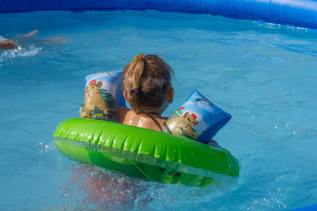 A little girl swims on an inflatable ring in the pool in summer. Back view of a child in the blue water of the pool. The child bathes on a hot summer sunny day.の写真素材