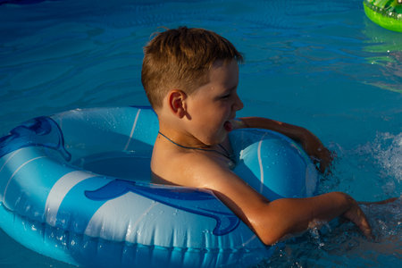 Boy child bathes in an inflatable pool on a blue circle in the summer in the backyard. In the rays of the setting sun, the child enjoys swimming in the pool in summer. Summer kids concept.の写真素材