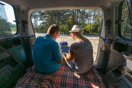 View from inside the car. Travelers sit in the trunk of a minivan and study the route on a tablet. Back view. A man and a woman in the forest take a break from traveling by car and study the tablet.の写真素材