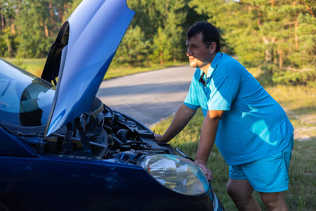 A handsome man stands in front of the open hood of a car that has broken down on the road and tries to fix it. During a family trip, the car breaks down and the man tries to fix it.の写真素材