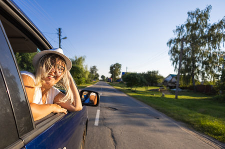 Leaning out the car window while driving, a woman smiles and looks into the camera. The wind beautifully develops the womans hair in the car window. The woman enjoys the feeling of freedom.の写真素材