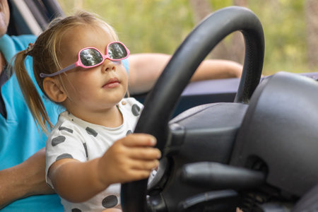 A cute little girl driving a car on her fathers lap. Child girl in sunglasses with a serious expression learns to steer a car. A father teaches his daughter how to drive a car.の写真素材