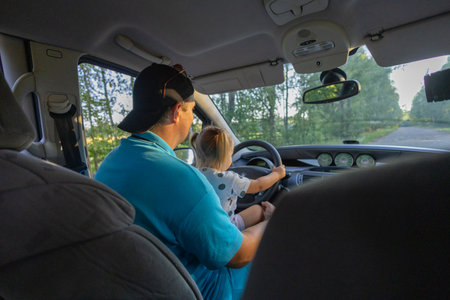 The father put the little daughter on his lap and let him drive under supervision. Father teaches daughter to drive. A little girl holds the steering wheel of a car while sitting on her fathers lap.の写真素材