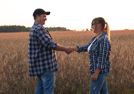 A beautiful couple of farmers at sunset inspects a field with wheat before selling grain Make notes on the tablet. Farmers smile and shake hands at sunset in the field. Agricultural business concept.の写真素材