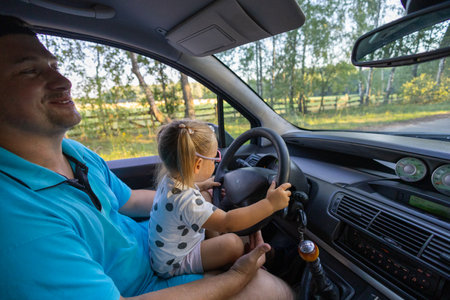 The father put the little daughter on his lap and let him drive under supervision. Father teaches daughter to drive. A little girl holds the steering wheel of a car while sitting on her fathers lap.の写真素材