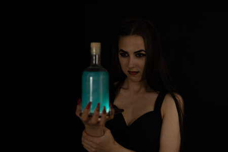 For Halloween, a beautiful witch brewed a potion. On a black background, a woman witch holds a bottle of poison in her hand. On a dark halloween night, a witch conjures over a potion.の写真素材