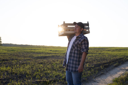 A tired worker carries a box of potatoes on his shoulder along the road along the field in the rays of the setting sun A man works in the field and harvests potato vegetables. End of the working day.の写真素材