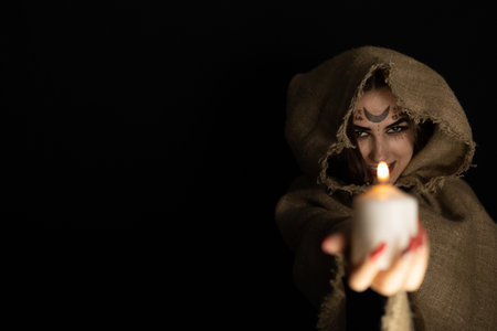 Copy space. On Halloween night, a hooded witch laughs and holds out a burning candle from the darkness. The witch conjures in the evening on halloween over the flame of a candle. Halloween concept.の写真素材
