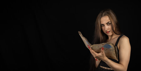 Copyspace. On a halloween night, a gothic woman is addicted to reading a book with black magic. A beautiful woman with long hair is holding a book with a text. Halloween and witchcraft concept.の写真素材