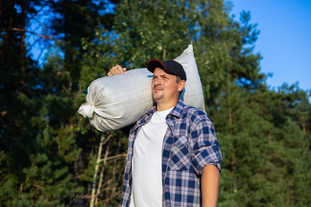 A man in a plaid shirt carries a sack over his shoulder from the field. Autumn harvest. A man harvests crops from the field. A farmer with a sack on his shoulder returns from the harvest.の写真素材