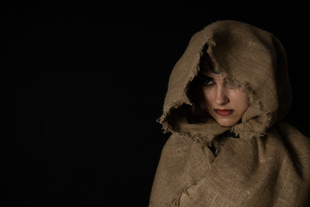In a cape with a hood, a beautiful female witch stands in the semi-darkness with runic symbols on her face. Halloween. Halloween black witch portrait. Halloween holiday concept.の写真素材