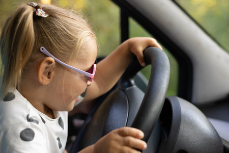 Close-up. Portrait. Funny little girl holding the steering wheel of a car and laughing. A small child cutely turns the steering wheel of a car while sitting on his fathers lap.の写真素材